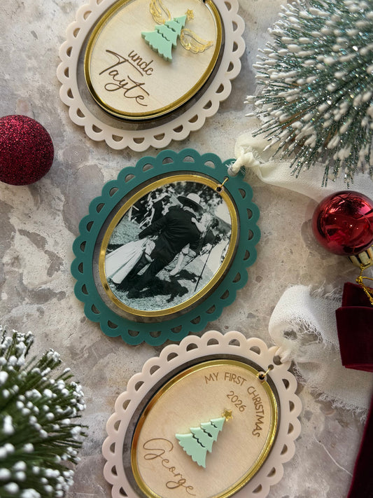 Three personalized oval Christmas ornaments with scalloped edges are arranged on a textured surface with holiday decorations. One ornament has a cream lace border and a mint green tree, reading 'uncle Tajte'.