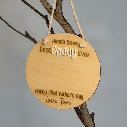 A round natural wood hanging plaque engraved with 'Hands Down Best Daddy Ever' and 'Happy First Father's Day Love Tom', suspended by a rope from a tree branch against a plain gray background.