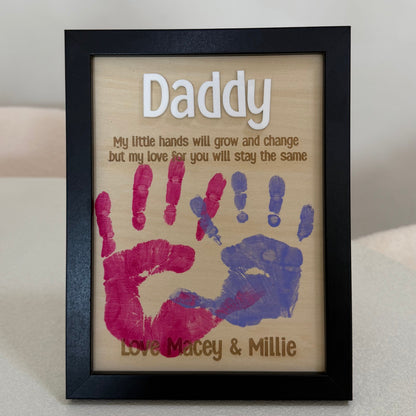 A personalized wooden framed plaque with a black frame, featuring the word 'Daddy' and a printed message, decorated with pink and blue handprints on a light wood background.