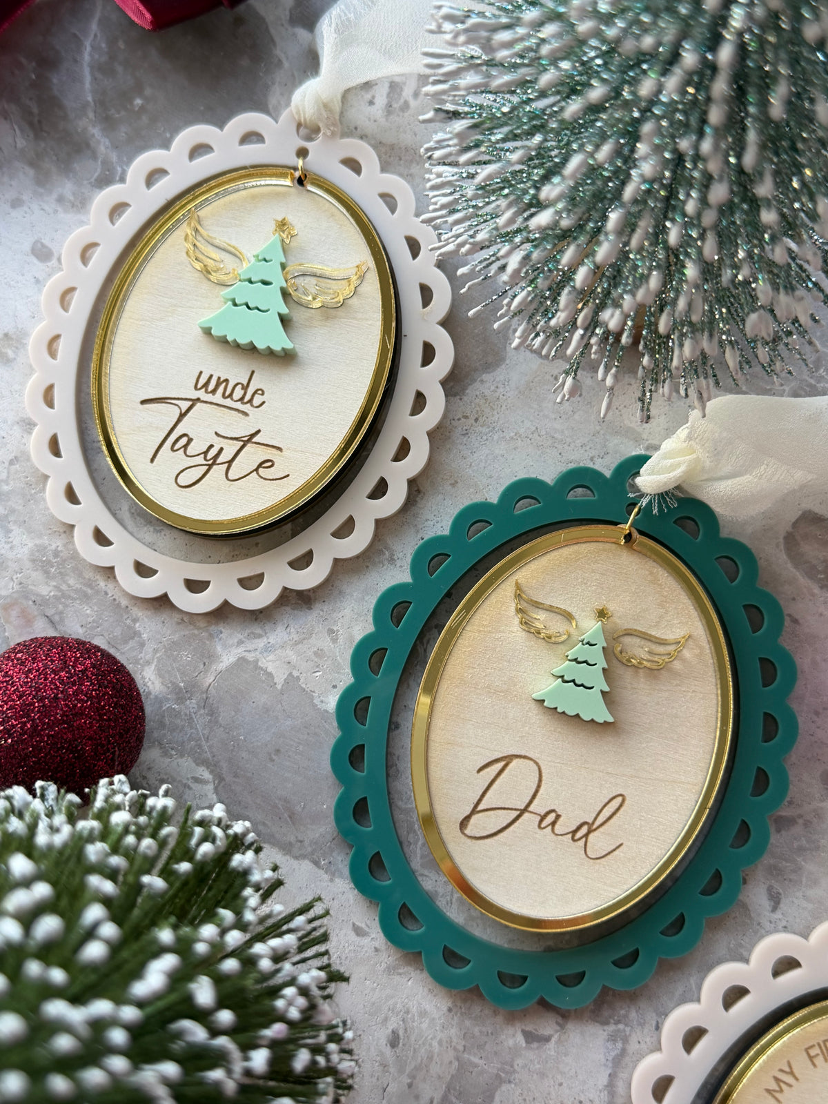 Two oval wooden Christmas ornaments with scalloped lace edges hang on a textured surface. One ornament is cream with a gold inner border and the name , featuring a mint green angel tree.