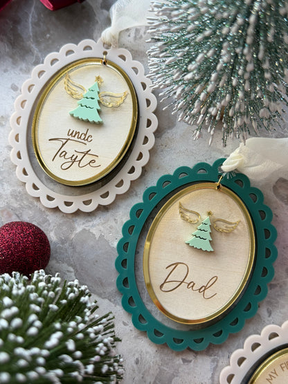 Two oval wooden Christmas ornaments with scalloped lace edges hang on a textured surface. One ornament is cream with a gold inner border and the name , featuring a mint green angel tree.