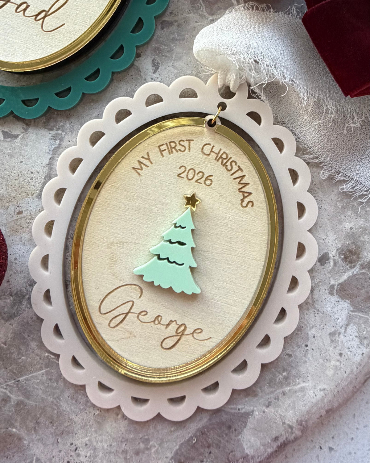 Personalized oval Christmas ornaments with scalloped edges, gold trim, and a mint green tree design. One ornament is engraved with 'My First Christmas 2026' and the name 'George', while another partially visible ornament in the background is engraved with 'Dad'.