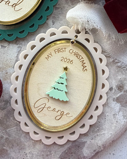 Personalized oval Christmas ornaments with scalloped edges, gold trim, and a mint green tree design. One ornament is engraved with 'My First Christmas 2026' and the name 'George', while another partially visible ornament in the background is engraved with 'Dad'.
